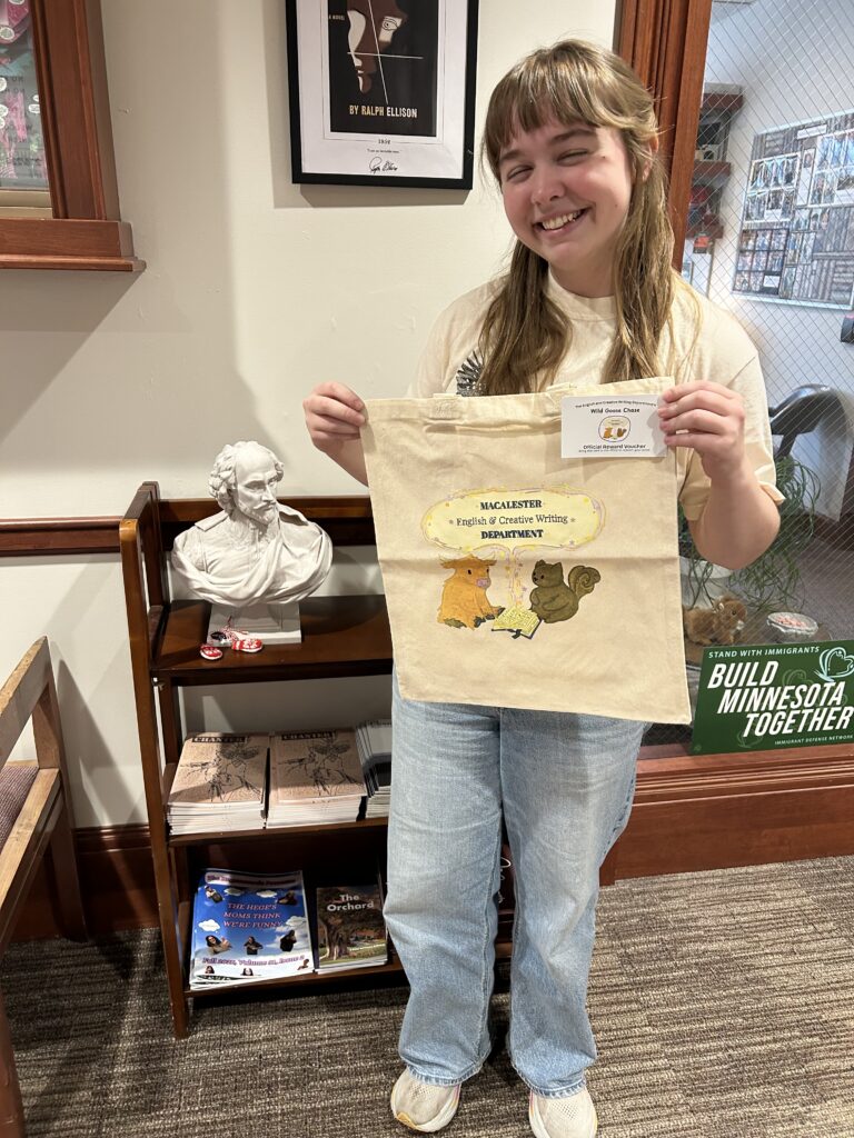 Photo of Sophia Woods '27 stands in front of a bust of Shakespeare and a collection of Chanter holding an English and Creative Writing Department tote bag.