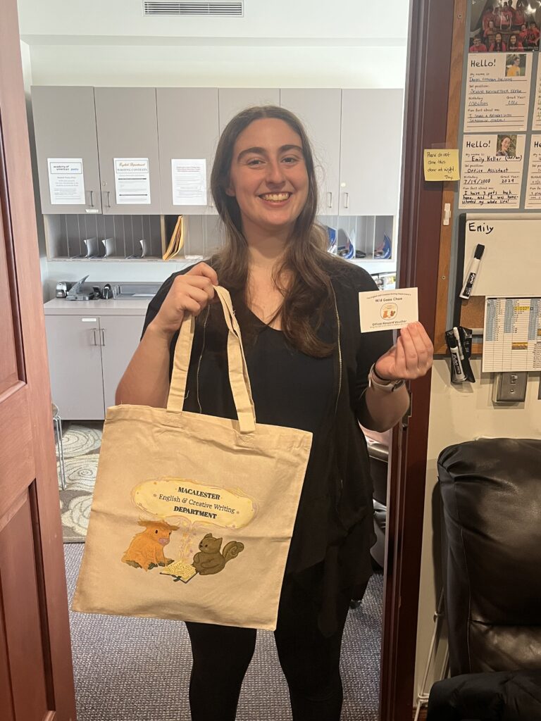 Natasha Friedman holds her a small voucher and tote bag with the English Department mascot, a highland cow and a squirrel, printed on them.