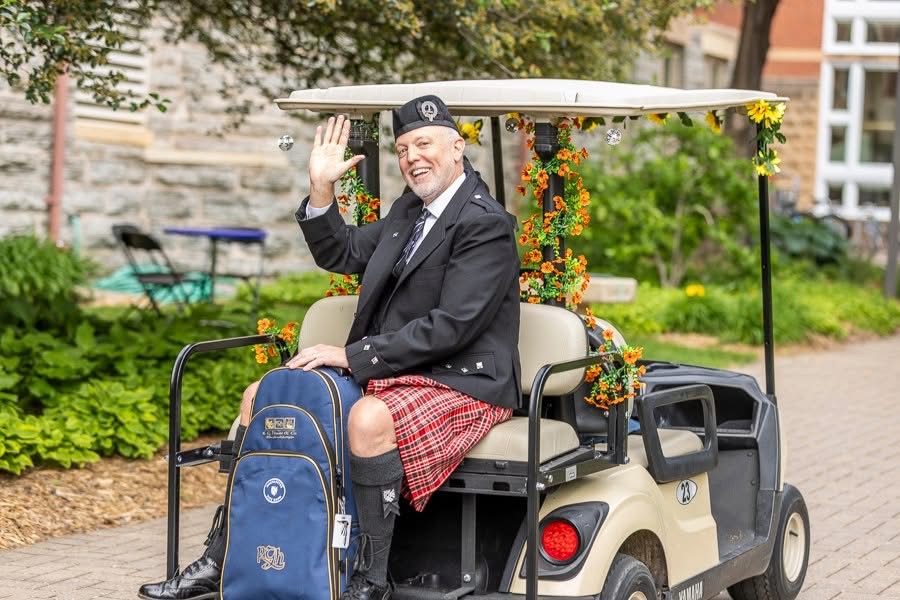 Photo of Mike Householder wearing a kilt and holding a bagpipe case on a golf cart outside Old Main