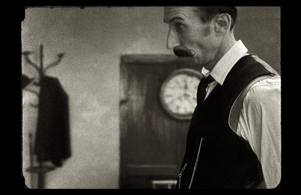 In a photo from the Acts of Memory Film, a man wearing a tie and formal attire stands before a clock.