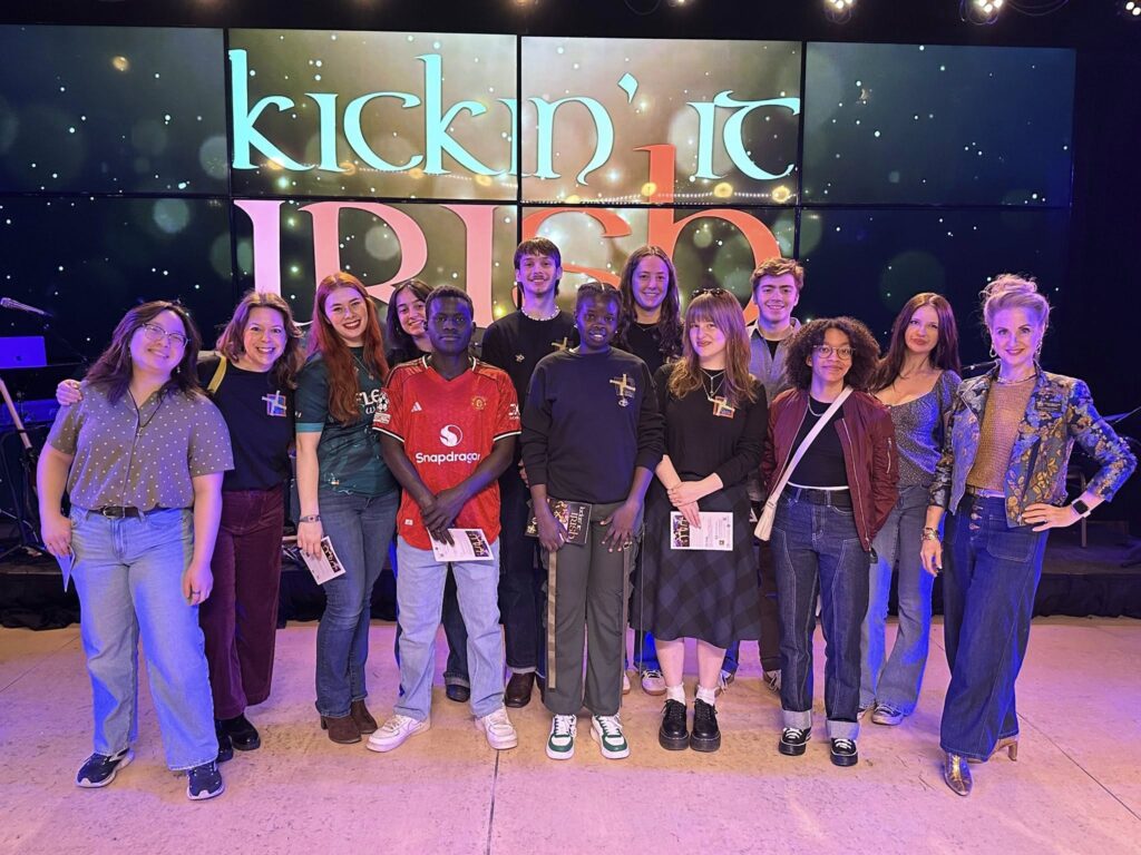Prof. Amy Elkins, students from her Irish Literature class and Executive Director of Celtic Junction Natalie Nugent O'Shea stand on stage before a brightly colored "Kickin' It Irish" background.