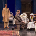 One actor stands while another kneels in front of a third, who is seated