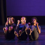 Three dancers sit on stage with their legs and arms off the ground