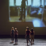 Several dancers stand turned away from the audience, watching a projection