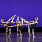 Dancers stand in a circle, holding hands, with legs lifted into the circle