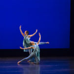 One dancer leans on the ground while another lifts their leg behind them, both have their arms reaching upward