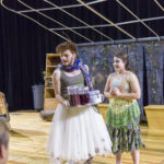 Two actors face the audience, one in a green skirt and one in a white skirt and holding a tray of glasses