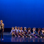 Several dancers sit in a line while another stands at the end