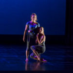 One dancer wraps their body around a second, standing dancer