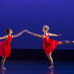 Two dancers in red dresses hold hands and lift their leg behind them