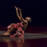 Dancer in red dress sits on stage with arms stretched upwards