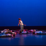 Several dancers lie around another, who stands
