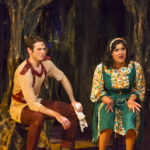 Two actors in colorful costumes sit in front of tree set pieces