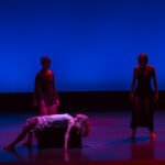 One dancer lies on a box and two stand in the shadows of the background
