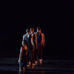 Four dancers stand in a line with their backs facing the audience