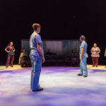 Several actors wearing scrubs stand all around the stage