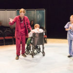 Two actors wearing scrubs stand next to a third, who sits in a wheel chair