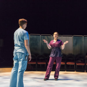 Two actors wearing scrubs face each other on stage