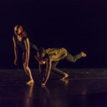 A dancer crouching on the floor grabs the leg of another, who leans forward