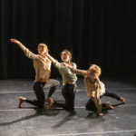Three dancers kneel on stage, reaching outward