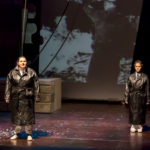 Two actors wearing black trench coats stand on stage
