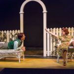 Two actors lounge on chairs in front of a white picket fence