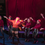Actors in red bellhop costumes surrounds another who is singing with their arms outstretched