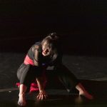 Dancer in black and red costume sits on stage