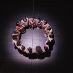 Several actors stand in a circle in the spotlight on stage