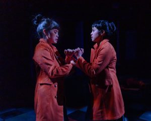 Two actors pinky-swearing on stage, wearing orange coats