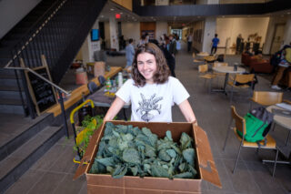 A student wearing a Mac Share t-shirt holds a large cardboard box filled with kale.