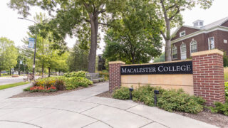 Macalester College's sign on Grand Ave. A sidewalk with a wooden bench leads people into campus next to the sign.