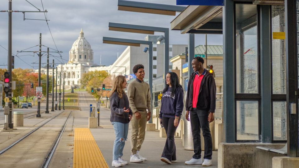 Four students chat while they wait at a Light Rail stop in front of the Minnesota State Capitol.