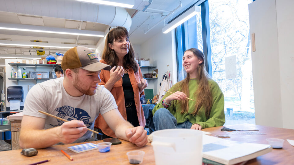 Two students holding paintbrushes talk with their professor.