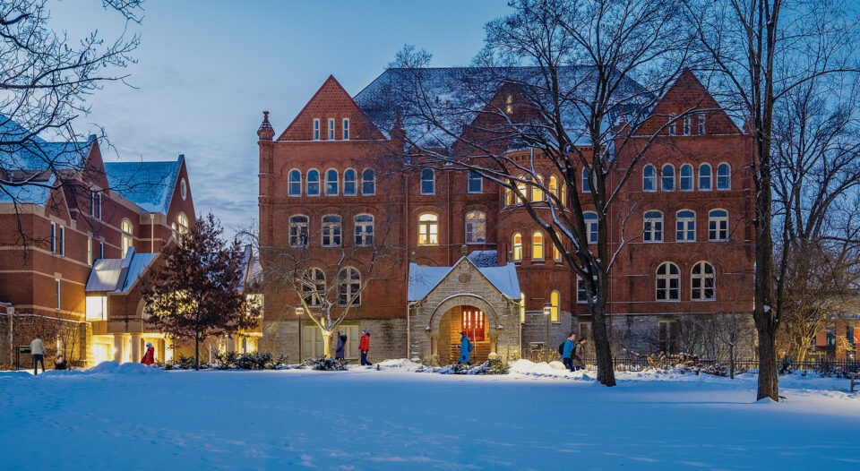 People walk in front of Old Main, a brick building with a large stone entrance, on a winter evening. Lights shine in the building's doors and windows.