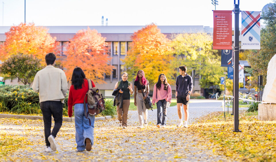 Students walk through campus. Red and orange fall leaves are visible on four trees behind them.