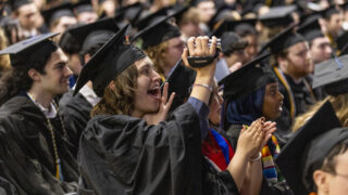 A crowd of students at Commencement. One is holding a camera pointed at the stage.