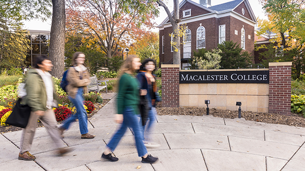 A group of 4 students walks past a brick sign that says Macalester College
