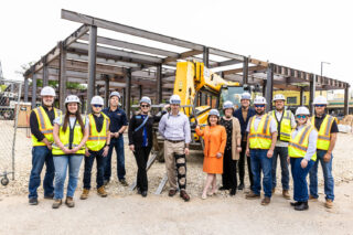 President Rivera and a group wear hard hats and stand in front of the under-construction residence hall and welcome center